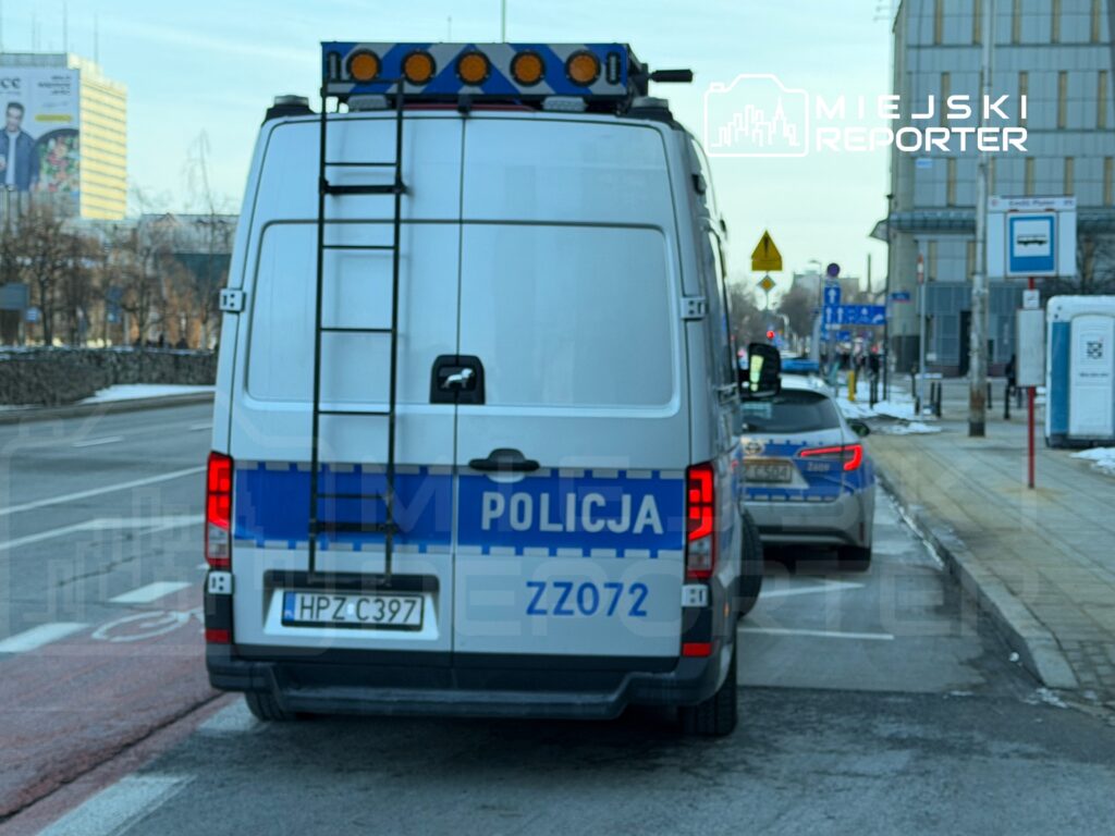 Policyjny van z włączonymi sygnałami świetlnymi zatrzymany na ulicy, obok nieoznakowanego radiowozu.