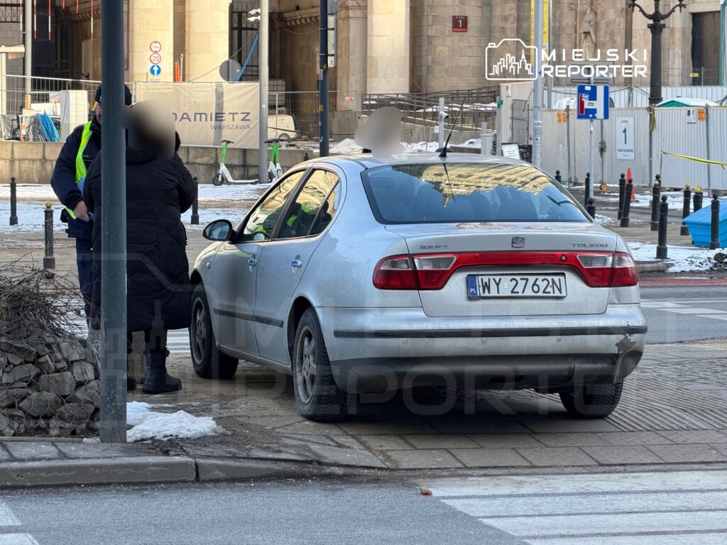 Kobieta w czarnym płaszczu rozmawia z policjantem obok zaparkowanego srebrnego samochodu.