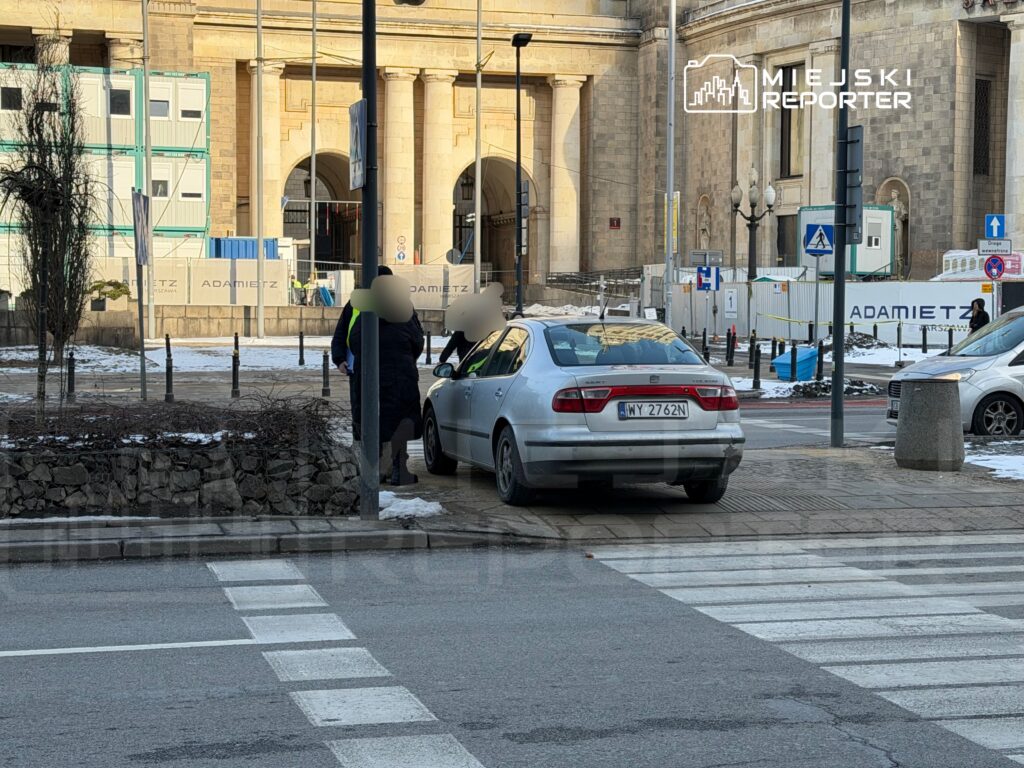 Mężczyzna w żółtej kamizelce rozmawia z osobą przy samochodzie zaparkowanym obok przejścia dla pieszych.