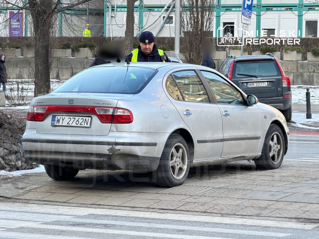 Mężczyzna w mundurze rozmawia z kierowcą przy samochodzie osobowym zaparkowanym na chodniku.