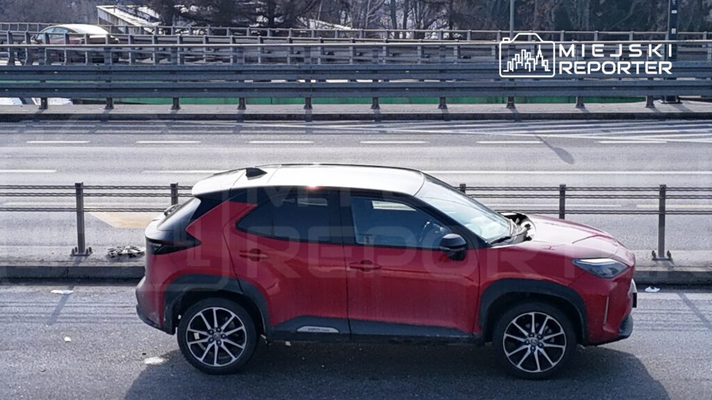 Czerwony SUV zaparkowany na poboczu ruchliwej drogi, obok metalowych barier ochronnych.