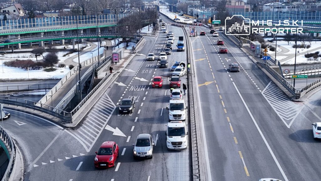 Ruch uliczny z samochodami osobowymi i dostawczymi na drodze, w tle widoczne zadrzewienie i budynki.