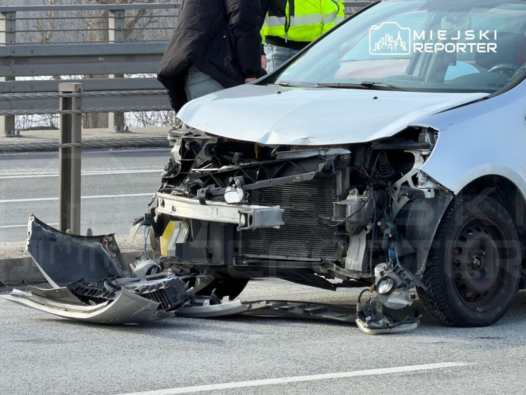Uszkodzony samochód osobowy z zniszczonym przodem stoi na poboczu drogi, obok dwóch osób w odblaskowych kamizelkach.