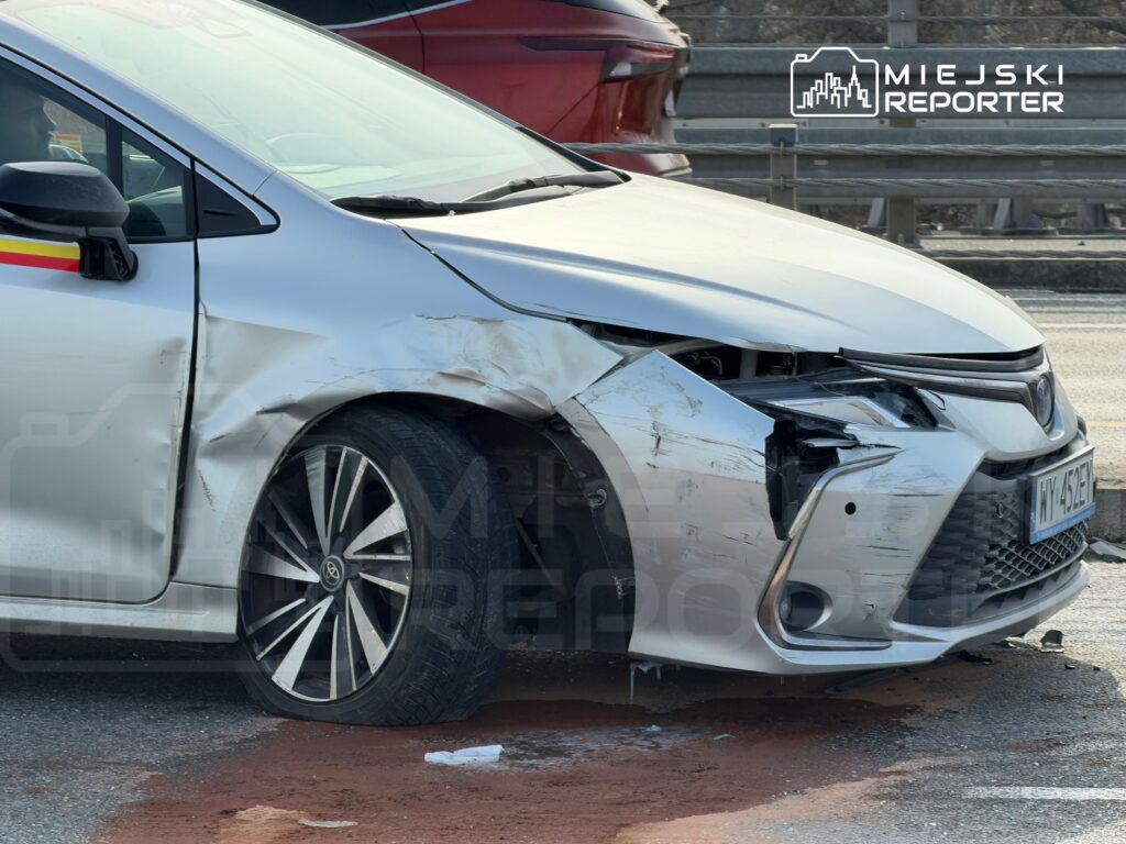 Uszkodzony samochód osobowy z wgniecioną przednią częścią stoi na poboczu drogi, obok widoczne inne pojazdy.