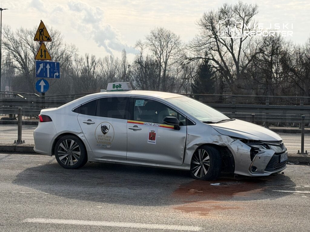 Uszkodzony samochód osobowy z logo Bolt stoi na poboczu drogi, obok znaków drogowych.