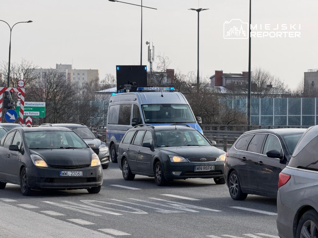 Policyjny radiowóz z włączonymi sygnałami świetlnymi porusza się wśród samochodów na ruchliwej drodze.
