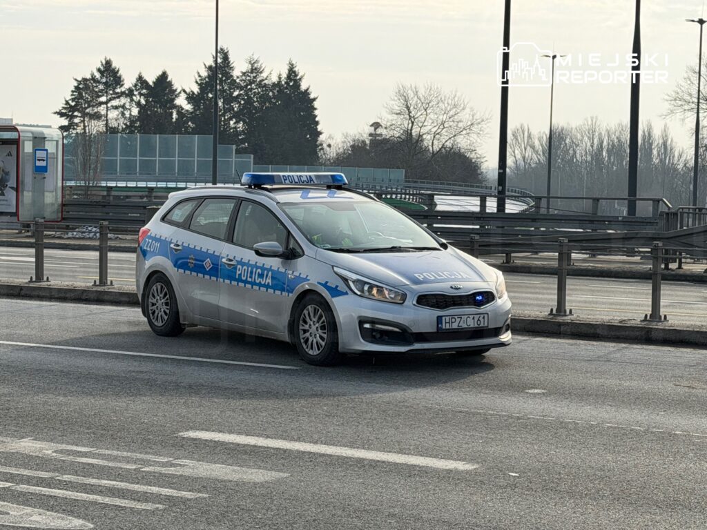 Policyjny radiowóz zaparkowany na pasie ruchu, z włączonymi sygnałami świetlnymi, w miejskim otoczeniu.