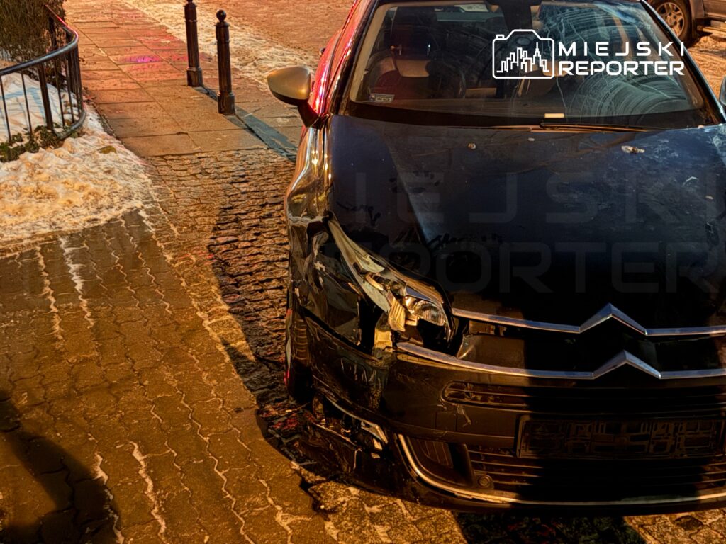 Czarny samochód osobowy z uszkodzeniami przodem stoi na oświetlonej ulicy, obok chodnika pokrytego śniegiem.