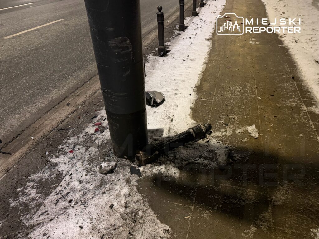 Złamany słup oświetleniowy leży na chodniku, wokół rozrzucone fragmenty szkła i śnieg.