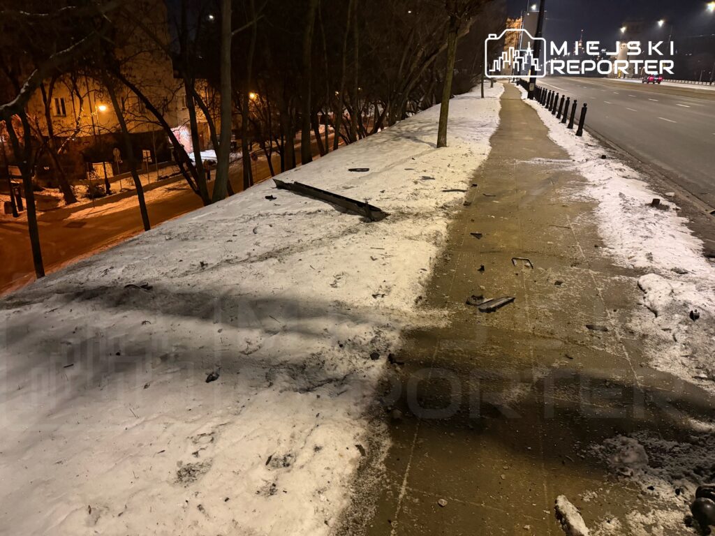 Zniszczony chodnik pokryty śniegiem, z porozrzucanymi fragmentami metalowymi, w tle oświetlona ulica nocą.