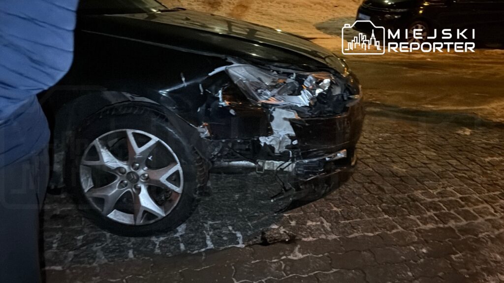 Czarny samochód osobowy z uszkodzoną przednią częścią stoi na oświetlonym parkingu.