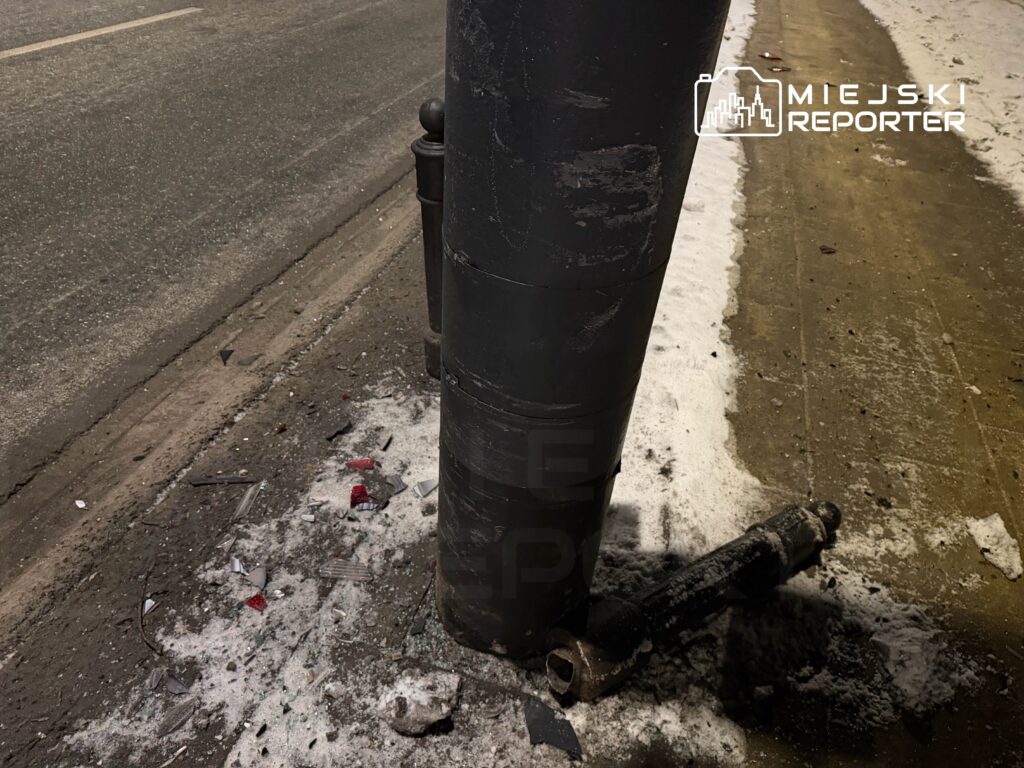 Złamana latarnia uliczna z uszkodzonym słupem i odłamkami szkła na chodniku w zimowym otoczeniu.