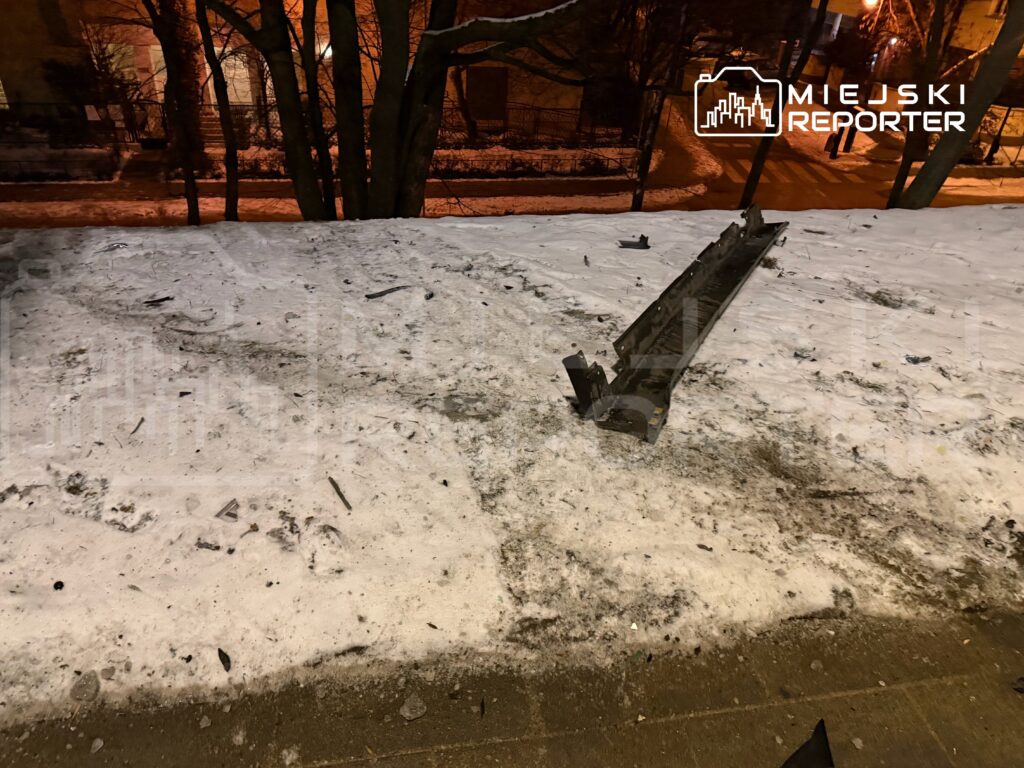 Na zaśnieżonej nawierzchni leży uszkodzona metalowa konstrukcja, wokół rozrzucone fragmenty. W tle widoczne drzewa i oświetlone budynki.
