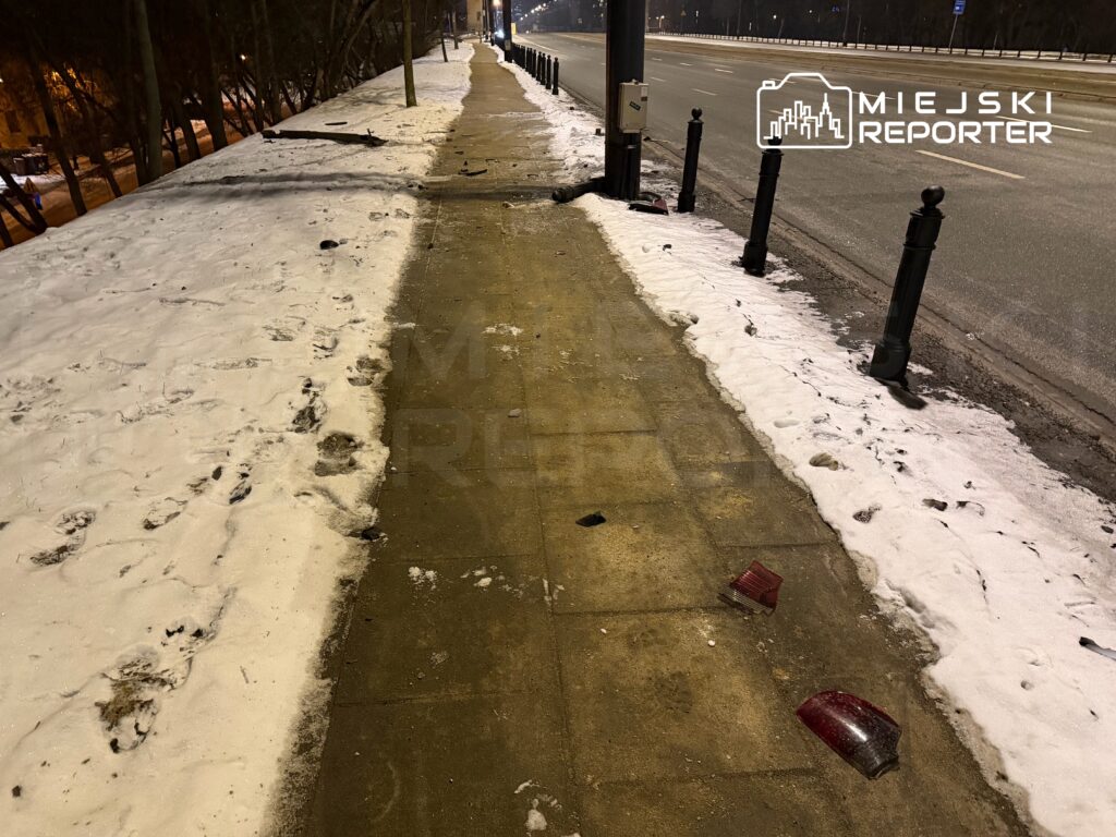 Na chodniku pokrytym śniegiem leżą fragmenty lamp samochodowych, obok biegnie pusta ulica.