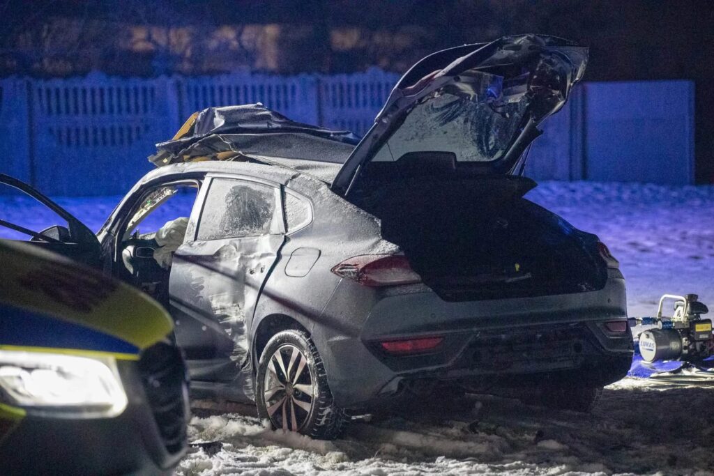 Uszkodzony samochód osobowy z otwartą klapą bagażnika stoi na ośnieżonej drodze, w tle niebieskie światła radiowozu.