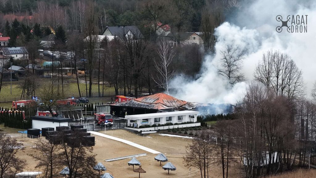 W tle widać budynek z płonącym dachem, z którego wydobywają się kłęby dymu, a w pobliżu pracują strażacy.