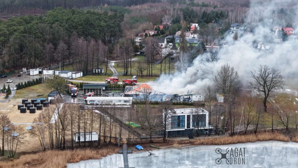 Pożar budynku w otoczeniu lasu, z widocznymi w tle wozami strażackimi i gęstym dymem unoszącym się w powietrzu.