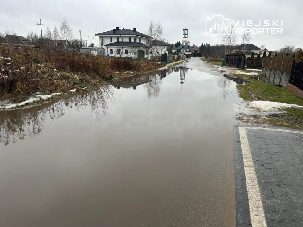 Zalany fragment ulicy z odbiciem budynków i drzew w wodzie, w tle widoczny dzwonnica i szary, pochmurny nieboskłon.