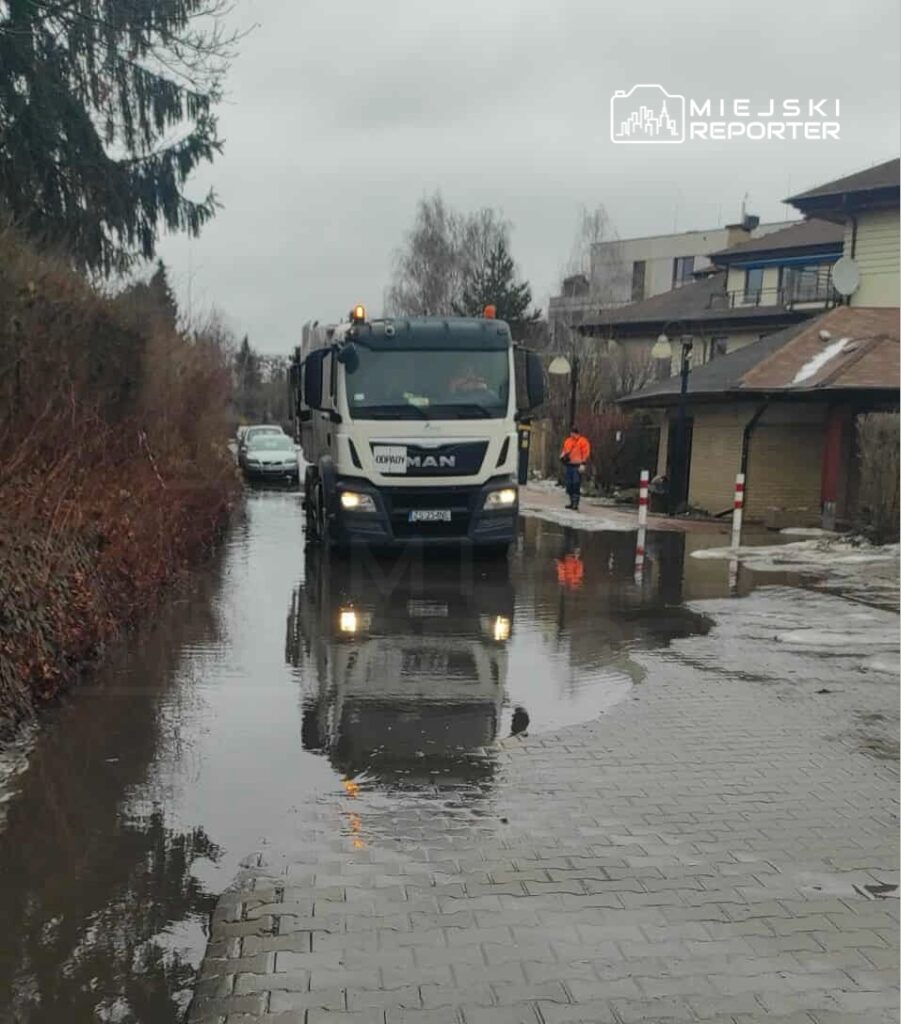 Ciężarówka MAN przejeżdża przez zalaną ulicę, obok której stoją dwa pracownicy w pomarańczowych kurtkach.