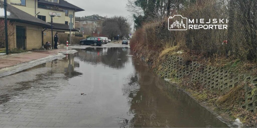 Zalany fragment ulicy z samochodami zaparkowanymi w tle oraz osobą stojącą obok budynku.