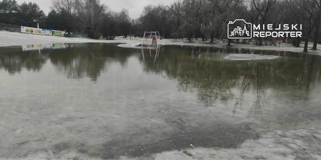 Zalany plac zabaw z huśtawką otoczony drzewami w deszczowy dzień. Woda odbija szare niebo.