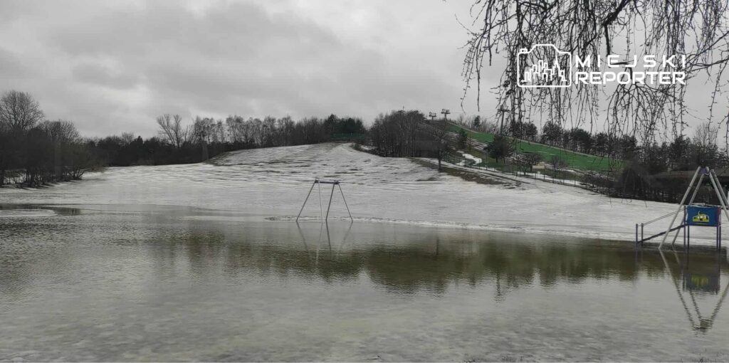 Zamoknięty plac zabaw z huśtawką w wodzie, otoczony śnieżnym krajobrazem i szarym niebem.