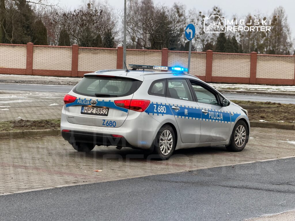 Radiowóz policyjny zaparkowany na poboczu, z włączonymi sygnałami świetlnymi, w otoczeniu trawnika i ogrodzenia.