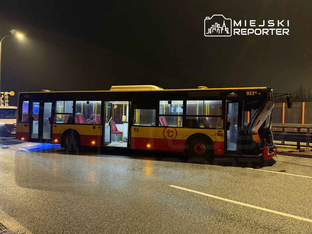 Autobus miejski z uszkodzoną tylną częścią stoi na mokrej nawierzchni drogi w nocy, obok zaparkowanego pojazdu.