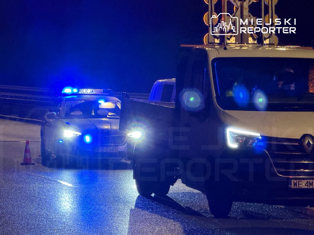 Policyjny radiowóz z włączonymi sygnalizatorami świetlnymi zatrzymuje pojazd na mokrej nawierzchni drogi.