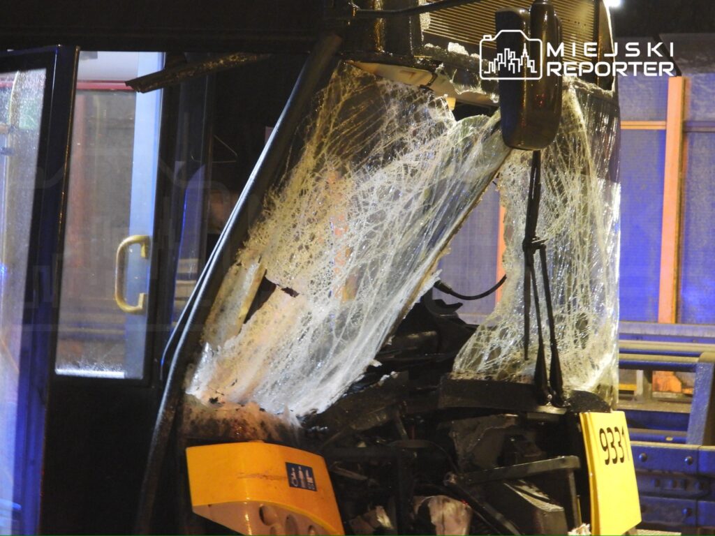 Uszkodzony przód autobusu miejskiego z rozbitą szybą i uszkodzeniami nadwozia, stojący na oświetlonej ulicy.