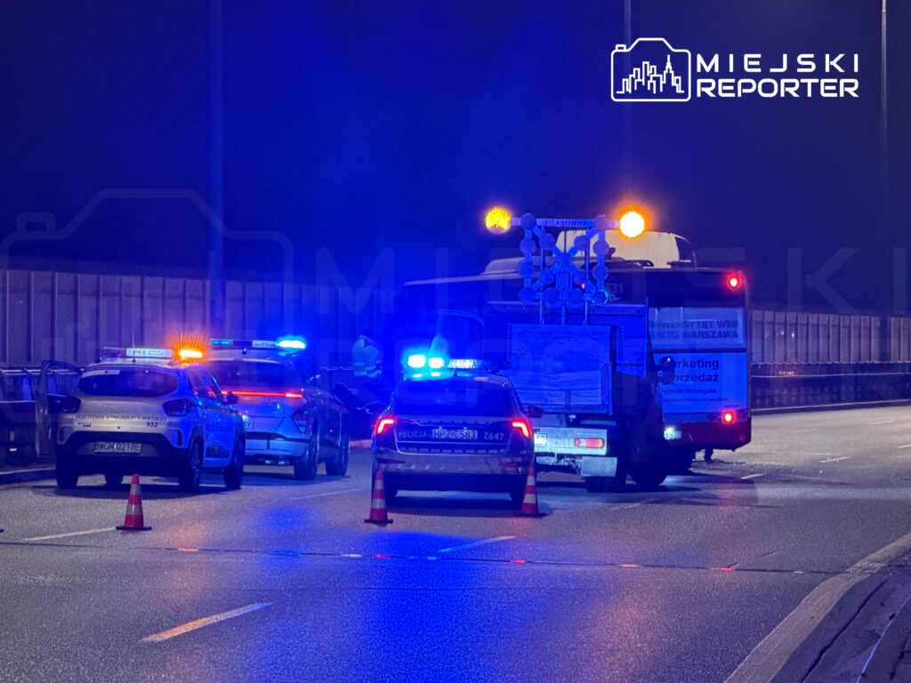 Na jezdni zatrzymane są trzy radiowozy policyjne oraz pojazd z sygnalizacją świetlną, otoczone pachołkami drogowymi.