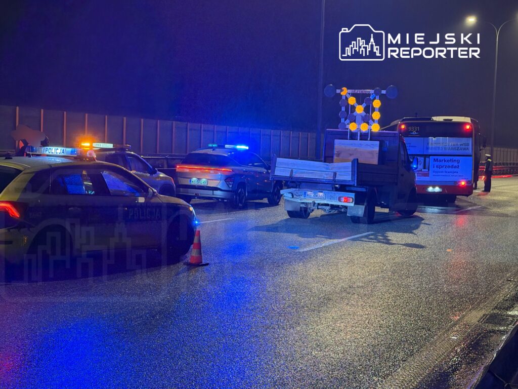 Policjanci w radiowozach z włączonymi sygnalizatorami zabezpieczają drogę, obok stoi ciężarówka z drewnem.