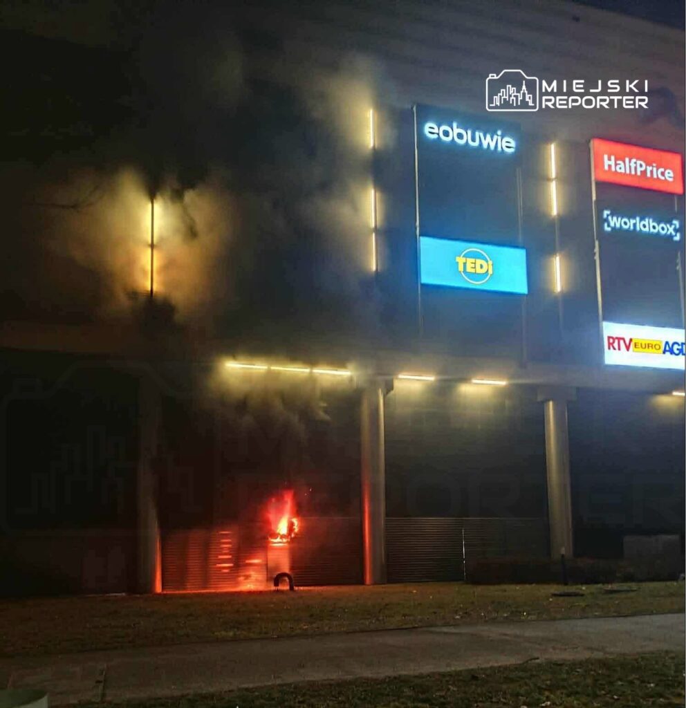 Ognisko pożaru widoczne na zewnętrznej ścianie nowoczesnego budynku handlowego, obok unoszące się kłęby dymu.