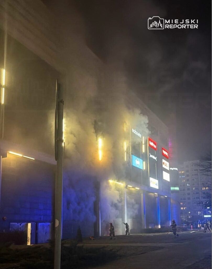 Dym wydobywający się z budynku handlowego, obok strażacy w akcji, nocna sceneria z oświetleniem neonowym.