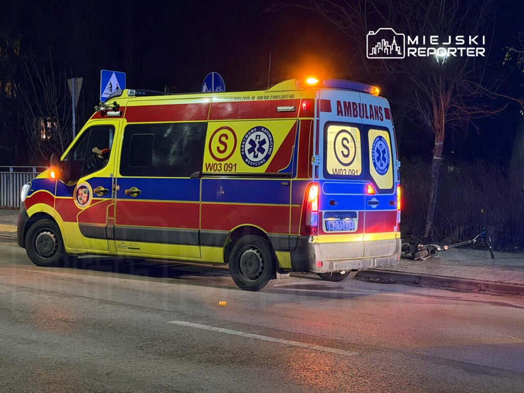Karetka pogotowia zatrzymana na ulicy w nocy, obok leżący rower. Światła awaryjne są włączone.