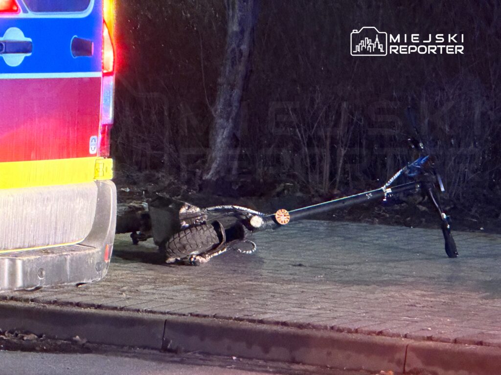 Złamany skuter leży na chodniku obok policyjnego radiowozu, w otoczeniu ciemnego terenu z drzewami.