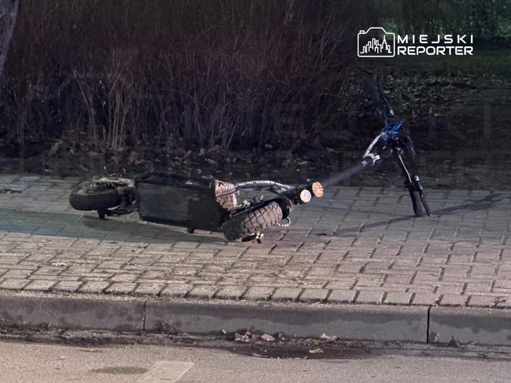 Uszkodzona hulajnoga leży na chodniku w zacienionym miejscu, obok krzaków.
