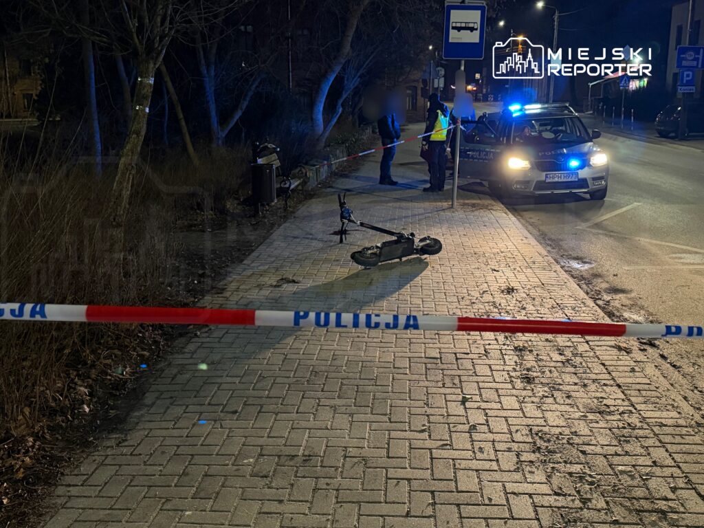 Na chodniku, przy policyjnej taśmie, leży uszkodzony skuter elektryczny, a w tle widać radiowóz i funkcjonariuszy.