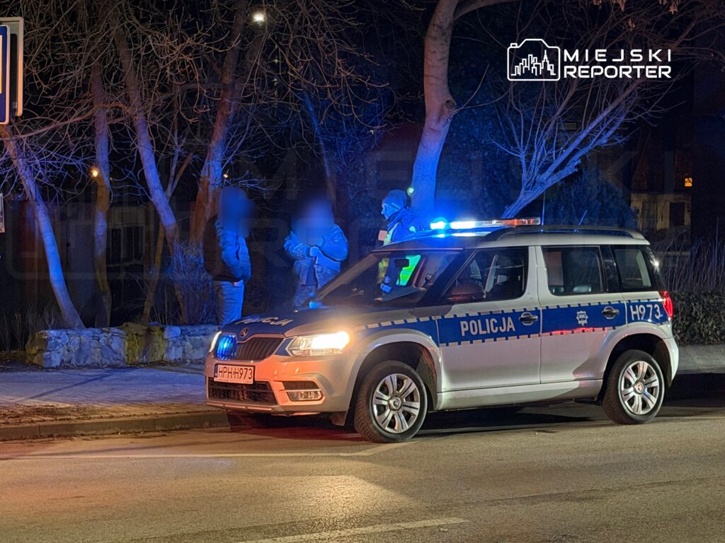 Policja rozmawia z dwiema osobami obok radiowozu na oświetlonej ulicy w nocy.