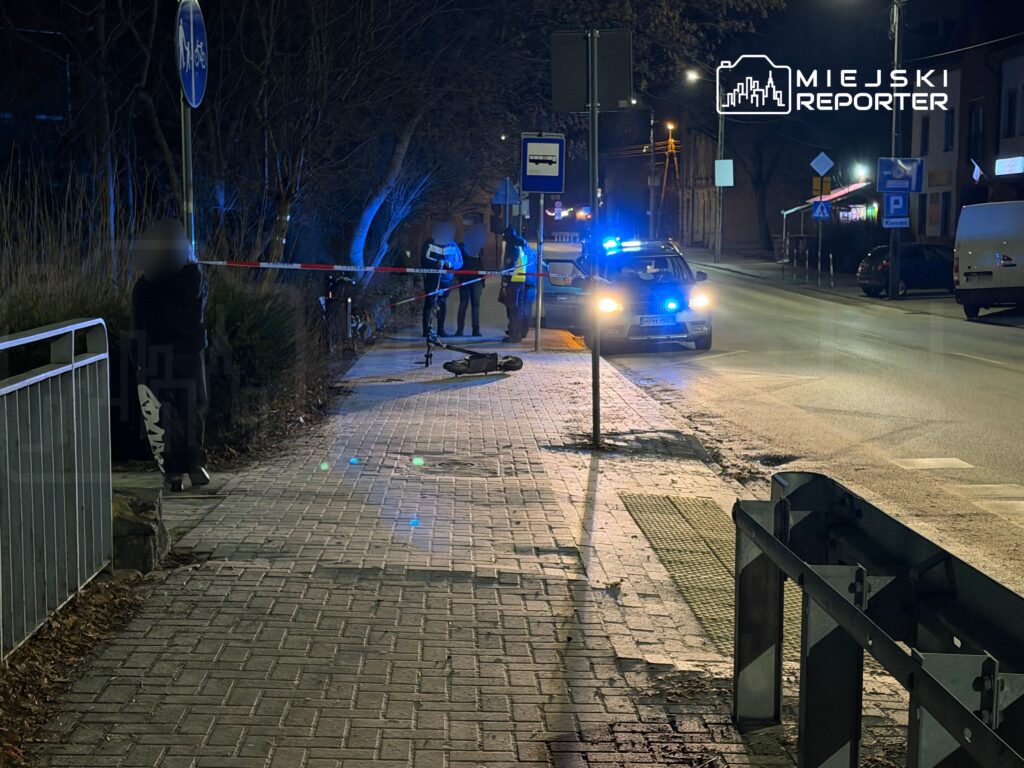 Grupa funkcjonariuszy policji zabezpiecza miejsce zdarzenia na oświetlonej ulicy, obok zaparkowanych pojazdów.