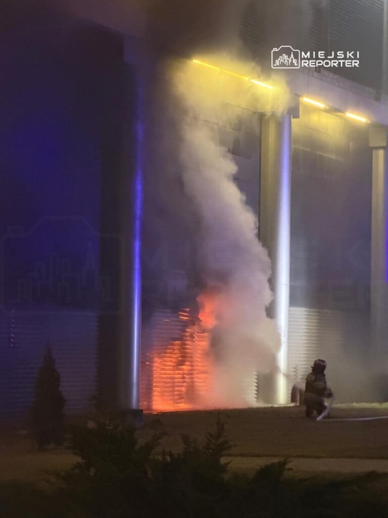 Strażak gaszący ogień w pobliżu budynku, z którego wydobywa się dym i płomienie.