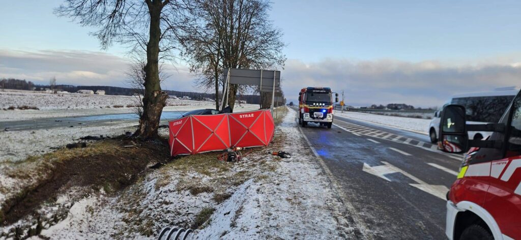 Na poboczu drogi stoi czerwona namiotowa osłona z napisem "STRAŻ", obok widać woz strażacki oraz leżące narzędzia.