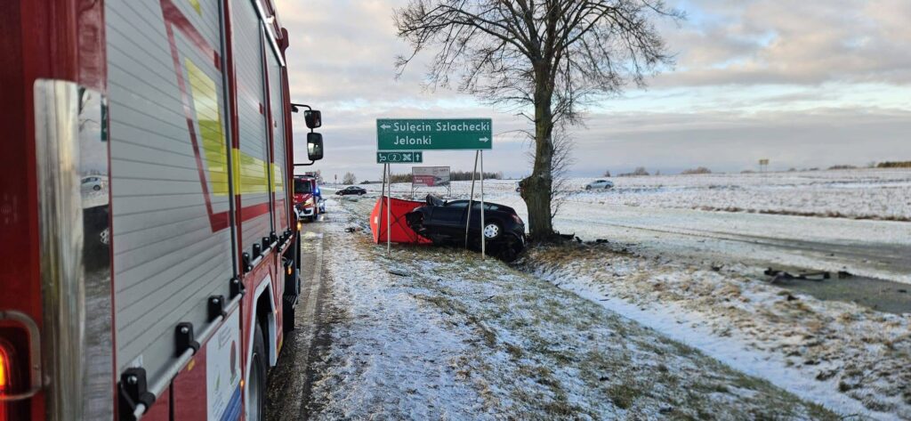 Czarny samochód osobowy w rowie, obok znaki kierunkowe z nazwami miejscowości, w tle wóz strażacki.