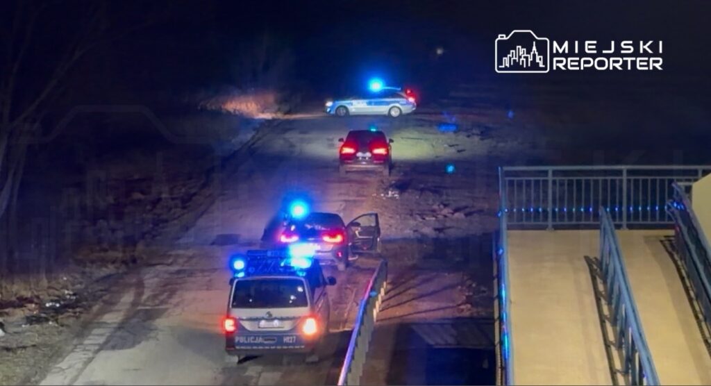 W nocy na opustoszałej drodze znajdują się dwa radiowozy policyjne z włączonymi niebieskimi światłami.