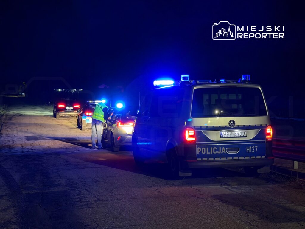 Policjant w odblaskowej kamizelce przeprowadza kontrolę drogową na opustoszałej drodze w nocy.