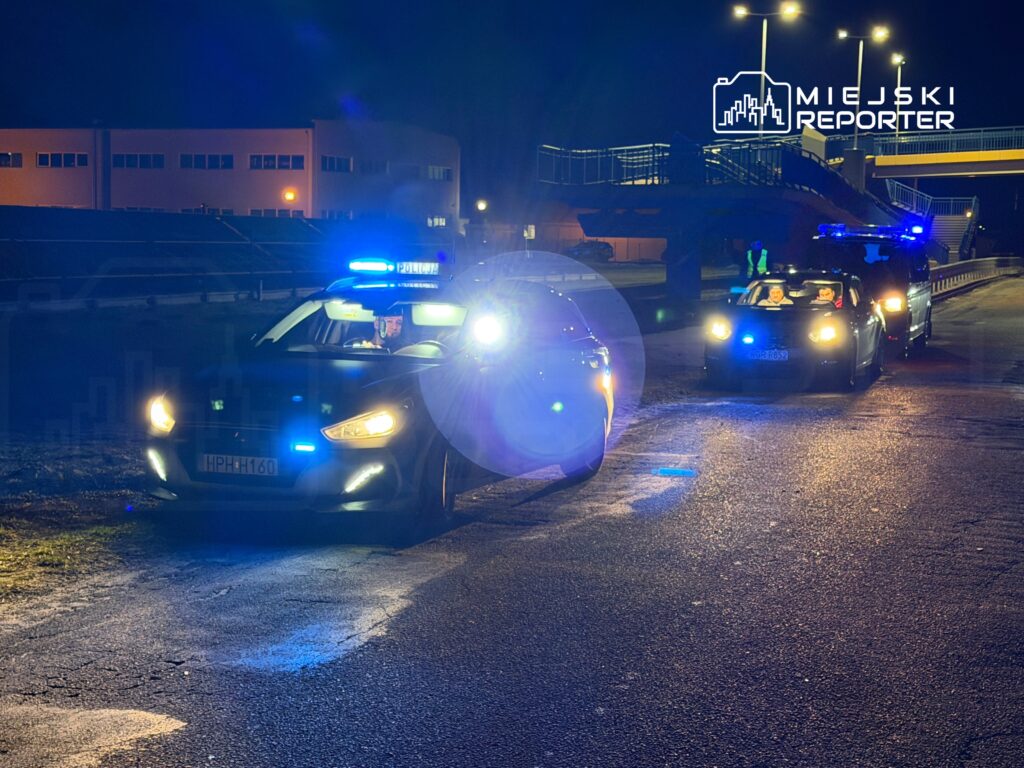 Policyjny radiowóz z włączonymi sygnałami świetlnymi stoi na drodze nocą, obok inny pojazd.