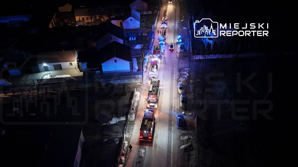 Na ulicy zaparkowane są pojazdy straży pożarnej i radiowozy, oświetlające okoliczne budynki w nocy.