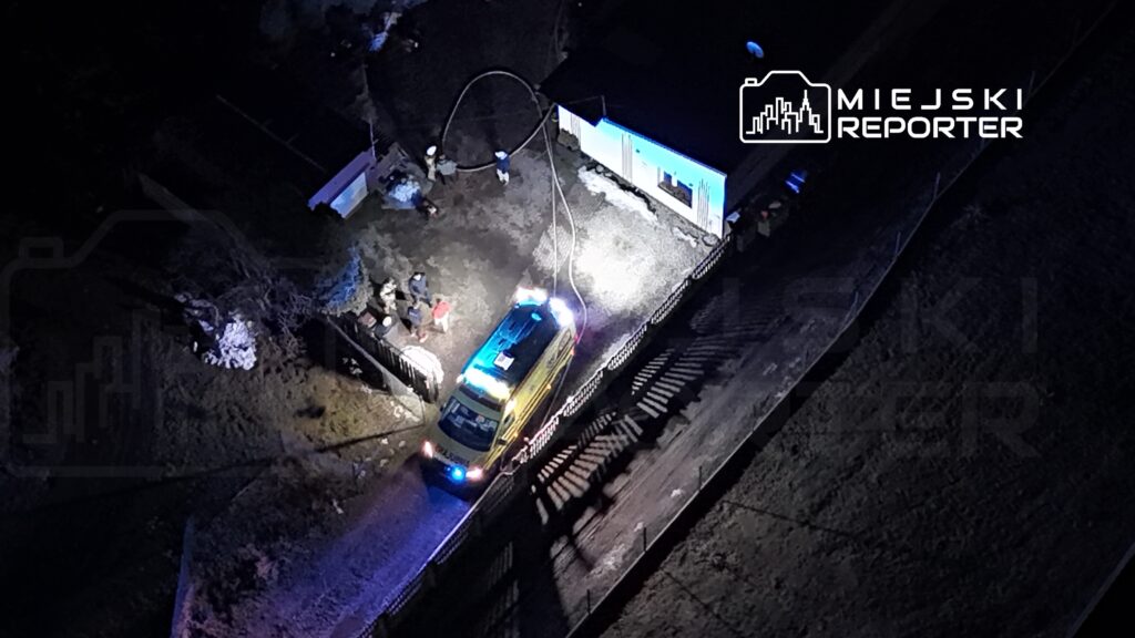 Zespół ratunkowy pracujący przy oświetlonym budynku, z karetka na drodze oraz osobami w pobliżu.