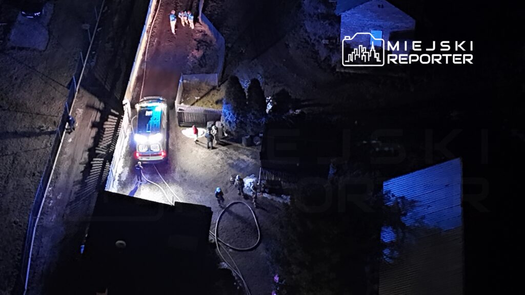 Zespół strażaków pracuje przy wozie strażackim na terenie posesji, podczas akcji ratunkowej w nocy.