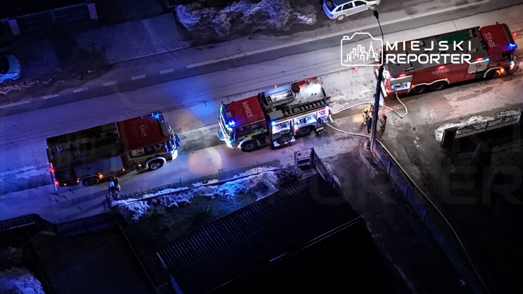 Dwa wozy strażackie z włączonymi sygnałami świetlnymi stoją na oświetlonej ulicy, a strażacy rozkładają węże.